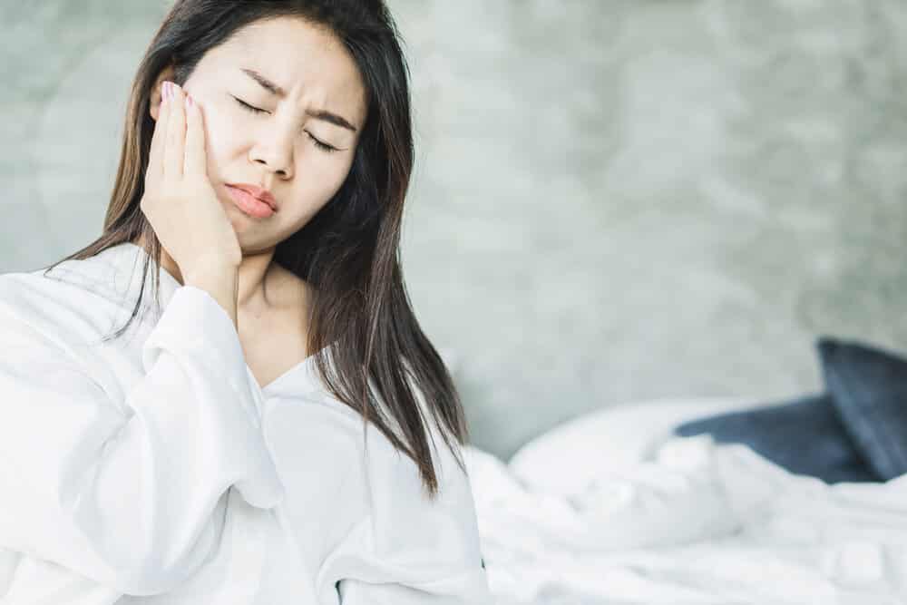 Asian woman suffering from gum disease, possibly gingivitis, holding hand to jaw and wincing in pain